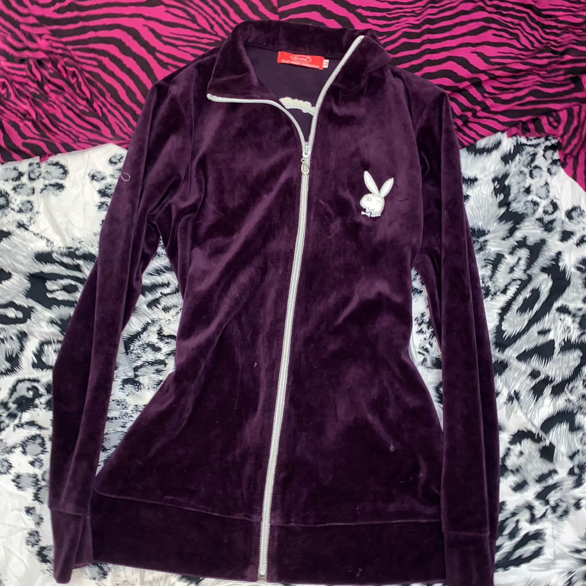 A dark purple velvet zip-up jacket with a white zipper and a white Playboy bunny logo on the chest, laid flat on a patterned surface.
